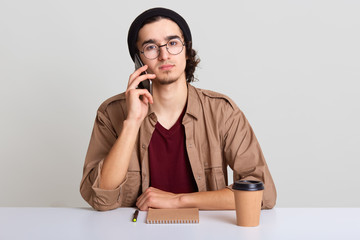 Photo of young man using smart phone for conversaion, discusses ideas for new starup, young hipster male student sitting at whte desk, drinking coffee, wearing casually. People and business concept.