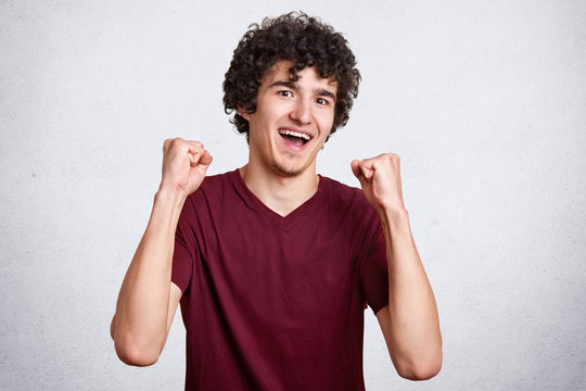 Studio shot of cheerful attractive European male being excited and glad to achieve victory, expresses positivity, clenches fists, screming hooray, feels proud, being champion and successful person.
