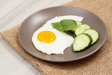 Plate with fried egg, fresh cut cucumber and leaf of basil on sackcloth.