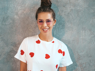 Naklejka na ściany i meble Closeup portrait of young beautiful smiling woman looking at camera. Trendy girl in casual summer white dress and sunglasses. Funny and positive female posing near gray wall in studio