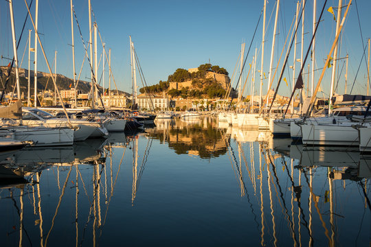 The castle of the Spanish port of Denia with the marina and many sailboats in the foreground with blue sky and sunshine with beautiful reflections in the water.