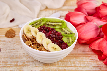 Bowl with Granola, banana, kiwi, berry with pink tulip flowers bouquet on rustic wooden table, breakfast on Mothers day or Womens day. Good morning. Copy space.