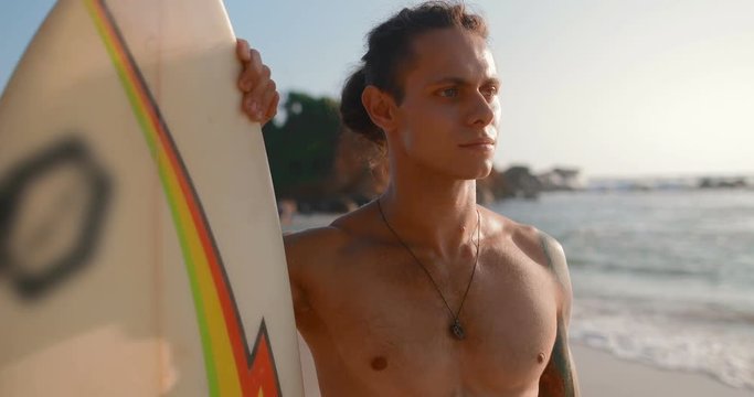 Portrait of a surfer full of strength and confidence. A guy stands with a board on the beach surrounded by palm trees.