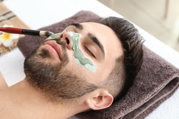 Cosmetologist applying clay mask onto face of young man in beauty salon
