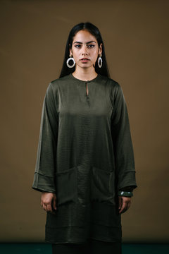 Portrait of a tall, slim and beautiful Indonesian Muslim woman in an olive green silk traditional dress (baju kurung) in a studio. She is standing elegantly against a brown background and posing