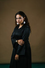 Studio portrait of a beautiful and attractive Malay Asian woman in a traditional black baju kurung dress against a brown background. She has frizzy beautiful hair and is posing for her portrait. 