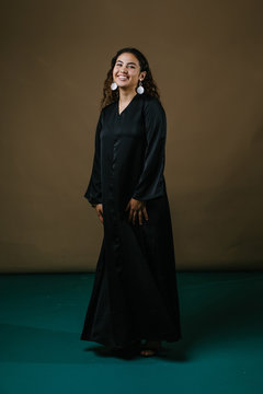 Studio portrait of a beautiful and attractive Malay Asian woman in a traditional black baju kurung dress against a brown background. She is dancing and smiling as she poses for her photo. 
