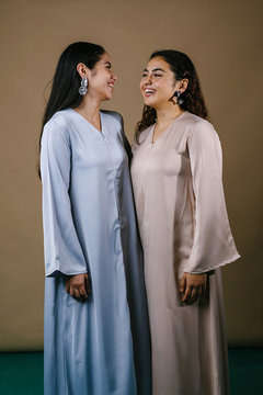 Portrait of two young Middle Eastern Muslim women smiling as they pose for their photo. They are wearing traditional pastel coloured Baju Kurung dresses to visit during Raya.
