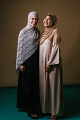 Two Muslim Middle Eastern sisters in traditional dresses and hijab head scarves stand and pose for a portrait together in a studio. They are both young, beautiful, elegant and attractive.