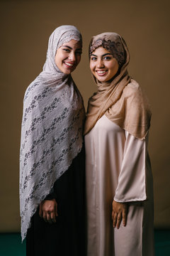 Two Muslim Middle Eastern sisters in traditional dresses and hijab head scarves stand and pose for a portrait together in a studio. They are both young, beautiful, elegant and attractive. 