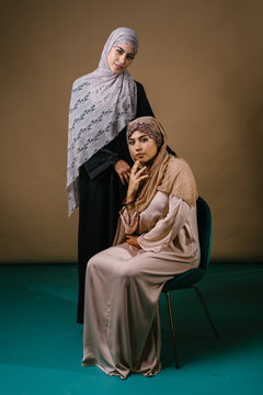 Two Indonesian Asian Muslim sisters in traditional dresses and hijab head scarves pose for a portrait together in a studio. They are both young, beautiful, elegant and dressed for Raya.