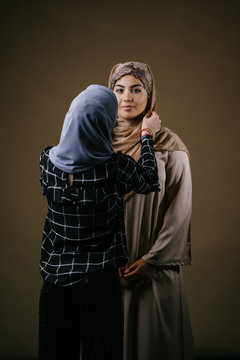A young and beautiful Muslim woman in a traditional outfit and hijab head scarf in a studio while her friend is adjusting her hijab. 