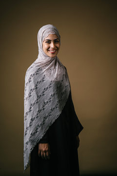 Studio portrait of a tall, slim, beautiful and elegant Middle Eastern Muslim woman in a black dress and hijab head scarf against a brown background. She is smiling happily in her fashionable outfit.