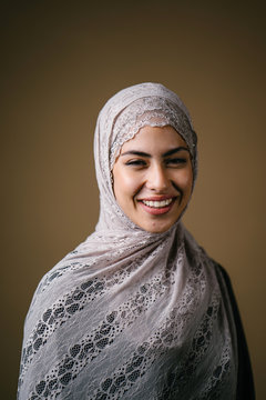 Close up studio portrait of a tall, slim, beautiful and elegant Middle Eastern Muslim woman in a black dress and hijab head scarf against a brown background. She is smiling happily.