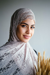 Close-up studio portrait of a tall, slim and elegant Middle Eastern Muslim woman in a black dress and hijab head scarf against a brown background. She is holding a bushel of wheat. 