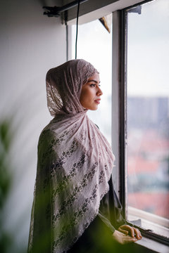Portrait of a beautiful and tall Malay Asian Muslim woman in a hijab head scarf standing by the window of her home. She is wearing a traditional outfit and is about have begin her fast. 