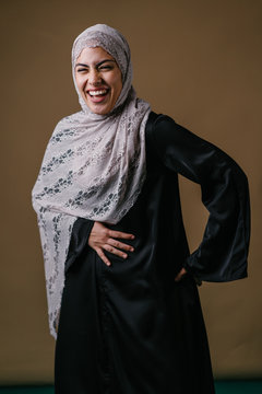 Portrait of a tall, slim and elegant Muslim Middle Eastern woman in a black dress and white hijab head scarf smiling and laughing as she poses for her head shot in a studio.