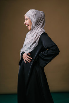 Studio portrait of a tall, slim, beautiful and elegant Middle Eastern Muslim woman in a black dress and hijab head scarf against a brown background. She is looking to the side and laughing.