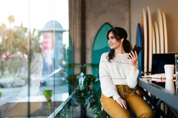 Young smiling woman sitting in cafe at table,looking out window, holding smartphone, listening to music on headphones. Lifestyle.