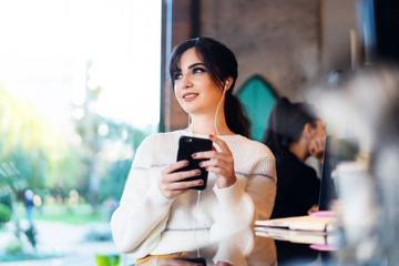 Young woman sitting cafe at table, thoughtfully looking out window, holding smartphone.Portrait girl with cell phone in her hands.