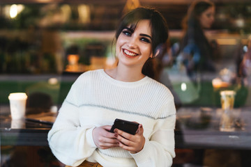 Portrait of smiling young woman with smartphone in her hands, sitting at table in cafe,looking at camera. Girl uses smartphone.