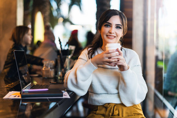 Portrait of smiling young woman with paper cup of coffee in her hands,sitting at table in cafe,looking at camera. Lifestyle.