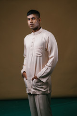 Studio portrait of a young, handsome and athletic Malaysian Indian Muslim Asian man in a cream colored traditional baju melayu tunic. He is dressed festively to go visiting for Raya.