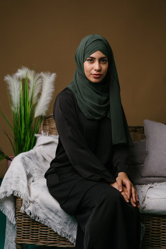 Portrait of a beautiful, elegant and attractive young Middle Eastern Muslim woman in a black dress and an olive green hijab head scarf. She has a neutral expression as she lounges on the couch at home