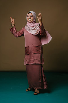 Portrait of a beautiful young woman in a traditional pink baju dress and hijab head scarf dancing in a studio. She is laughing and having fun.