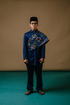 Studio Portrait Of A Happy, Handsome And Young Malay Asian Man In A Blue Baju Melayu Hat With A Patterned Sash Dressed For Hari Raya. He Is Ready To Go Visiting Friends And Family During Raya.