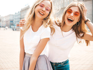 Fototapeta na wymiar Portrait of two young beautiful blond smiling hipster girls in trendy summer white t-shirt clothes. Sexy carefree women posing on street background. Positive models having fun in sunglasses.Hugging