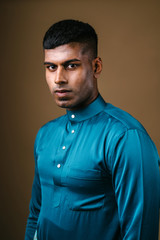Studio portrait of a young, handsome and athletic Malaysian Indian Muslim Asian man in a blue traditional baju melayu tunic. He is standing in front of a brown background in a studio.