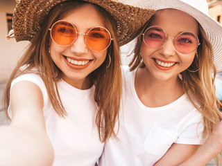 Two young smiling hipster blond women in summer white t-shirt clothes. Girls taking selfie self...