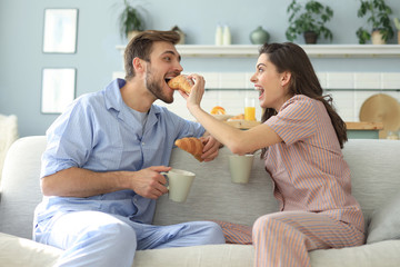 Obraz premium Happy young couple in pajamas in kitchen having breakfast, feeding each other a croissant.