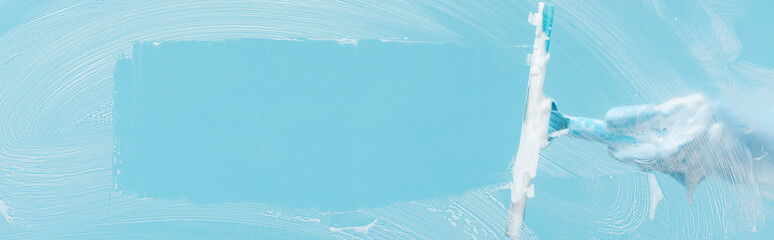 panoramic shot of woman in rubber glove cleaning glass with squeegee on blue background