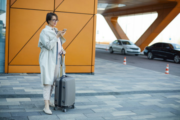 Serious stylish businesswoman in coat holding handle of wheeled luggage and using smartphone while waiting for taxi in airport area