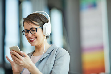 Excited attractive middle-aged woman in wired headphones using modern smartphone while chatting...