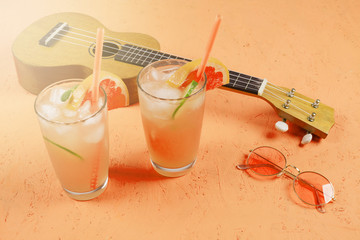 Glasses of citrus juice with ice cubes; sunglasses and ukulele on an orange textured backdrop