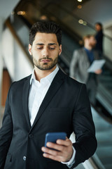 Serious handsome young mixed race entrepreneur in black suit standing in airport and using smartphone while corresponding through mobile app