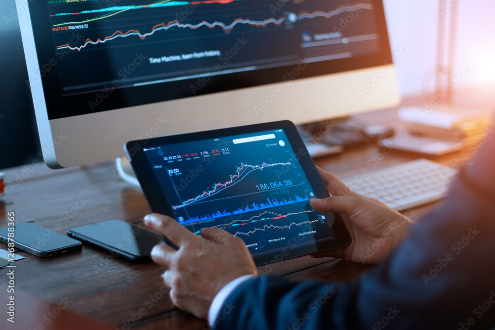 Poster Businessman checking stock market on digital tablet and a desktop computer with stock exchange graph on screen. Financial stock market. Analyzing data in office background.