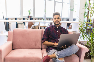Casual young indian man using laptop with happy on sofa in modern office