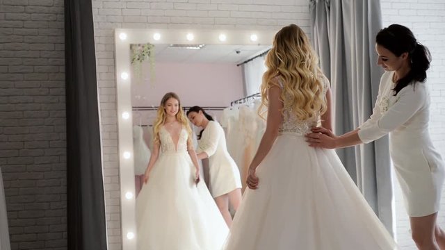 Wedding consultant helps bride choosen wedding dress in salon and wearing it near large mirror
