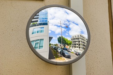 Outdoor convex safety mirror hanging on wall with reflection of an urban roadside view of cars...