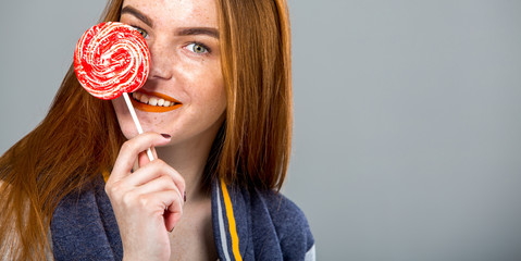 Beautiful woman with lollipop having fun. Playful red hair girl with long hair over grey background 