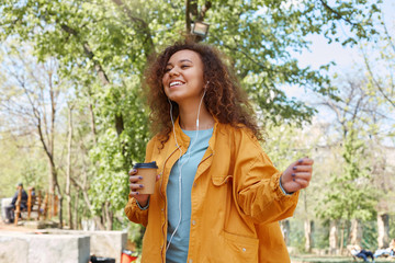 Portrait of young attractive dark skinned curly girl broadly smiling, holding a cup of coffee, wearing a yellow jacket, listening to music, walking in the park and enjoying the weather.