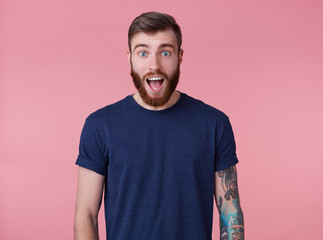 Shocked attractive red-bearded young guy with blue eyes, wearing a blue t-shirt, looking at the...