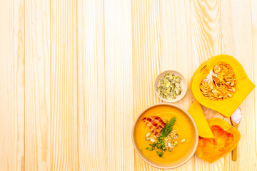 Homemade autumn hot pumpkin cream soup with smocked bacon and seeds. Raw pumpkin, fresh chives, dill, salt in spoon on wooden background, top view copy space.