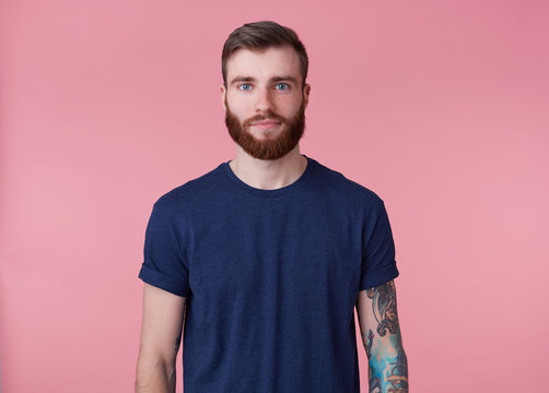 Young attractive red-bearded guy with blue eyes, wearing a blue t-shirt, looking at the camera with a calm facial expression isolated over pink background.