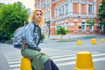 young attractive curly blonde woman sitting on a concrete fence near the pedestrian crossing