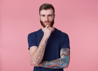 Young thinking attractive red-bearded guy with blue eyes, wearing a blue t-shirt, holds his hand at...